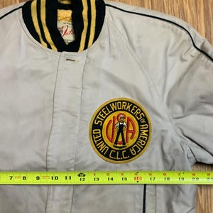 May include: A beige bomber jacket with a black and yellow striped collar. The jacket has a large embroidered patch on the left chest that reads "United Steel Workers of America, C.L.C.".