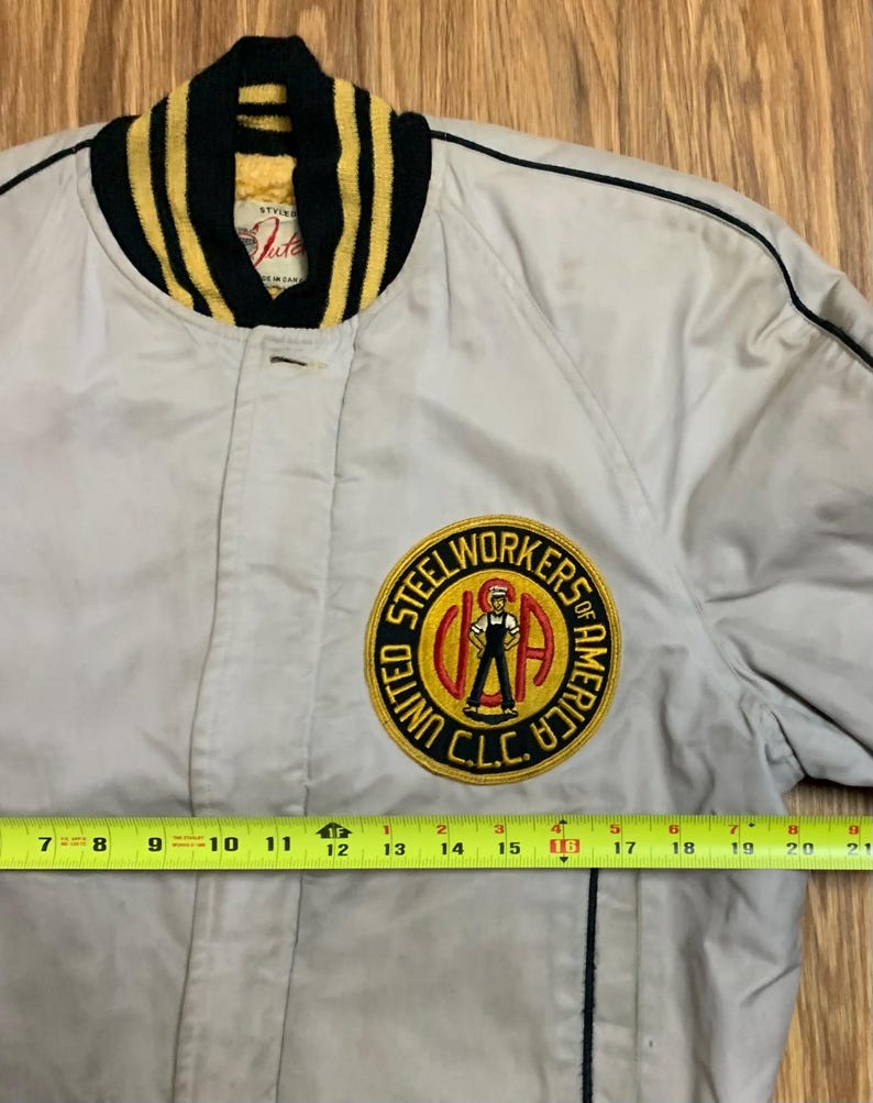 May include: A beige bomber jacket with a black and yellow striped collar. The jacket has a large embroidered patch on the left chest that reads "United Steel Workers of America, C.L.C.".