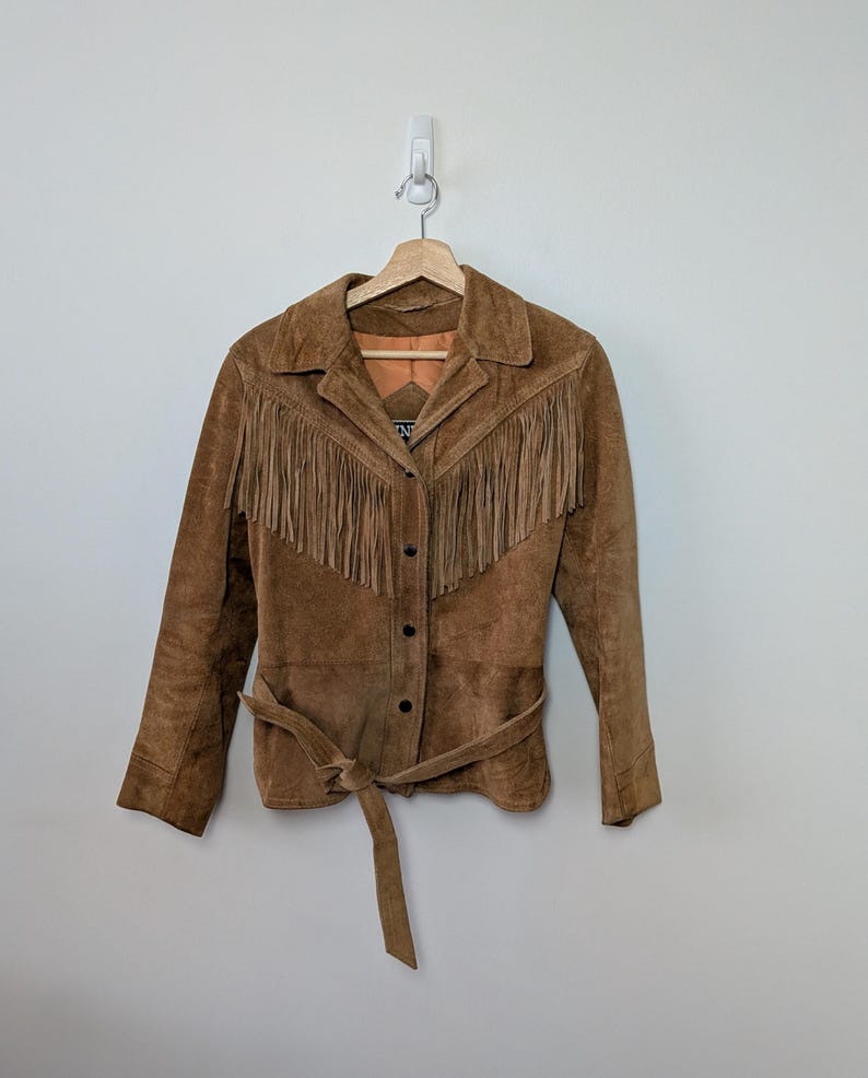 May include: A brown suede jacket featuring long fringe detailing across the chest and shoulders. The jacket includes a pointed collar, a front button closure with dark buttons, long sleeves, and a matching tie belt at the waist. The interior is lined.