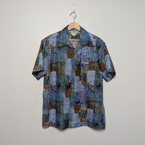 May include: Short-sleeved button-down shirt with a patchwork design in shades of blue, brown, and green. The shirt is hanging on a wooden hanger against a plain white wall. The fabric has a subtle sheen.