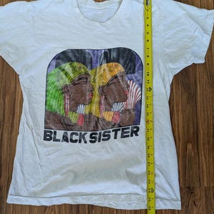 May include: White t-shirt featuring a graphic of two women with headscarves, one green and one yellow, with the text "BLACK SISTER" below. The shirt measures approximately 28 inches in length.