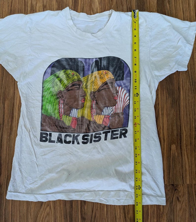 May include: White t-shirt featuring a graphic of two women with headscarves, one green and one yellow, with the text "BLACK SISTER" below. The shirt measures approximately 28 inches in length.