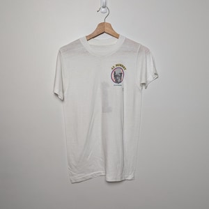 Puede incluir: Camiseta blanca con un pequeño gráfico en la parte delantera. El gráfico presenta un retrato de un hombre dentro de un círculo rosa con las palabras "AL BUMBRY" y "Baltimore" alrededor. La camiseta está colgada en una percha de madera.