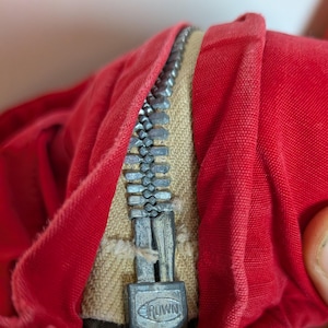 May include: Close-up of red fabric with a silver zipper. The zipper is partially open, revealing a beige fabric underneath. The zipper pull has the word "CROWN" on it.