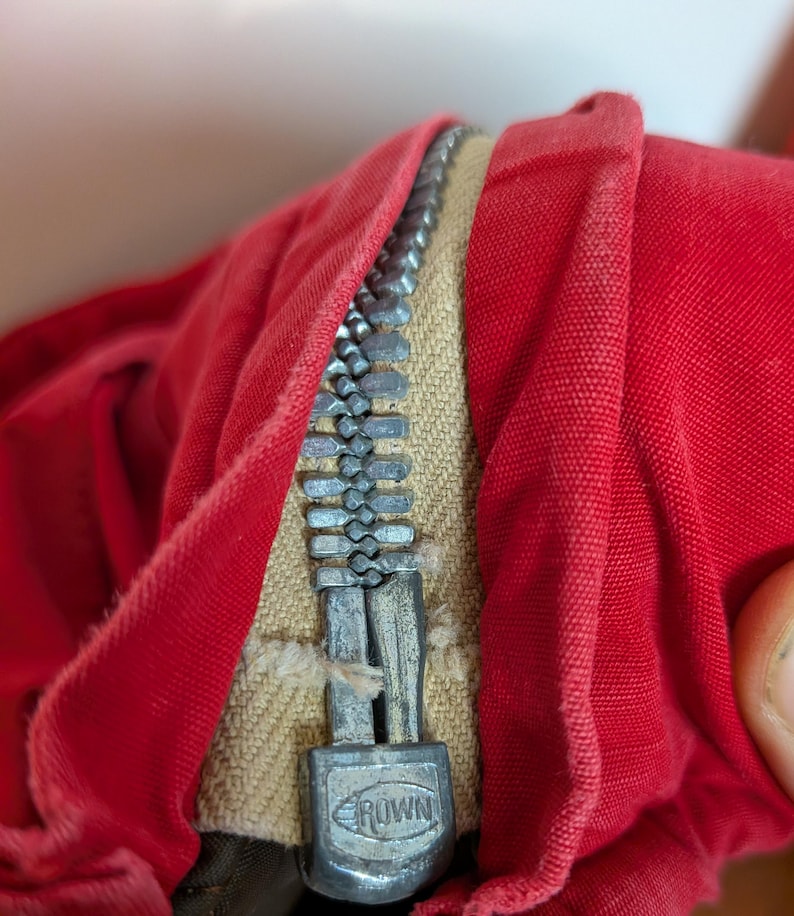 May include: Close-up of red fabric with a silver zipper. The zipper is partially open, revealing a beige fabric underneath. The zipper pull has the word "CROWN" on it.