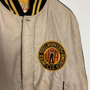 May include: A beige bomber jacket with black and gold striped ribbing around the collar and cuffs. The jacket has a patch on the left chest that says "Steelworkers of America, United C.L.C."