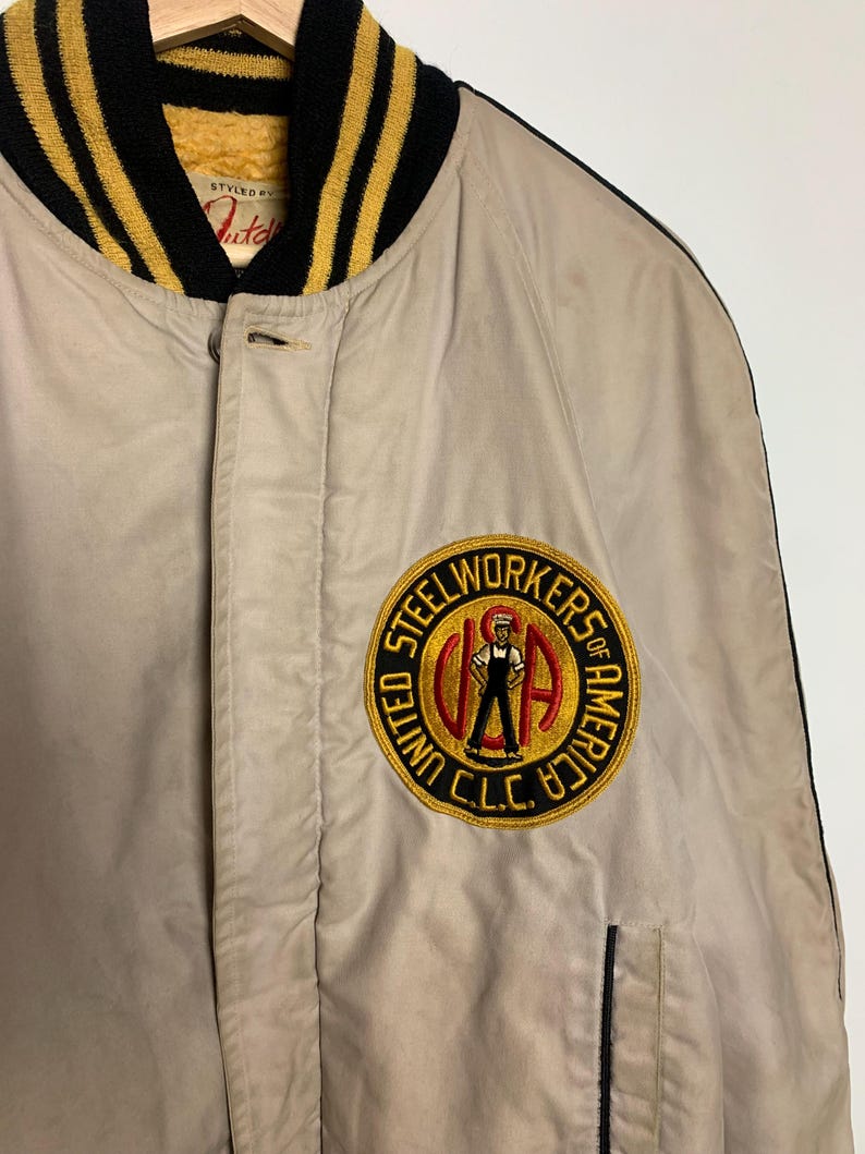 May include: A beige bomber jacket with black and gold striped ribbing around the collar and cuffs. The jacket has a patch on the left chest that says "Steelworkers of America, United C.L.C."