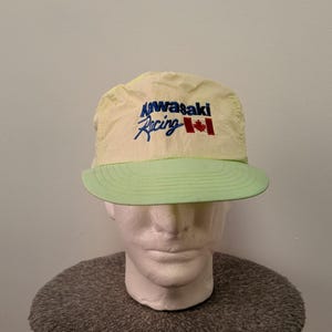May include: A pale yellow baseball cap with a light green brim. The front features embroidered text that reads Kawasaki Racing in blue and black, accompanied by a small red and white Canadian flag emblem on the right side of the text.