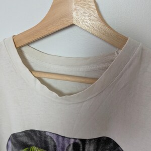 May include: Off-white t-shirt with a graphic print of a reptile with green and yellow scales, and red eyes. The shirt is hanging on a wooden hanger against a white background. The shirt's collar is visible.