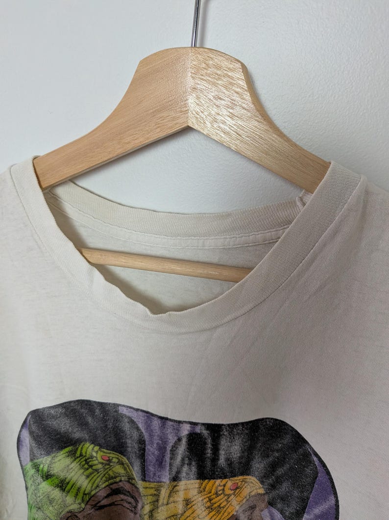May include: Off-white t-shirt with a graphic print of a reptile with green and yellow scales, and red eyes. The shirt is hanging on a wooden hanger against a white background. The shirt's collar is visible.