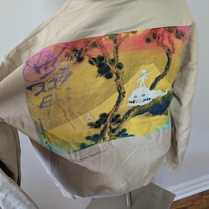 May include: A beige hooded sweatshirt featuring a large rectangular graphic on the back. The print displays a stylized landscape with yellow, red, and green tones, a white creature, and purple text. The text "Takashi Murakami / Kaikai Kiki Co., Ltd." is visible.