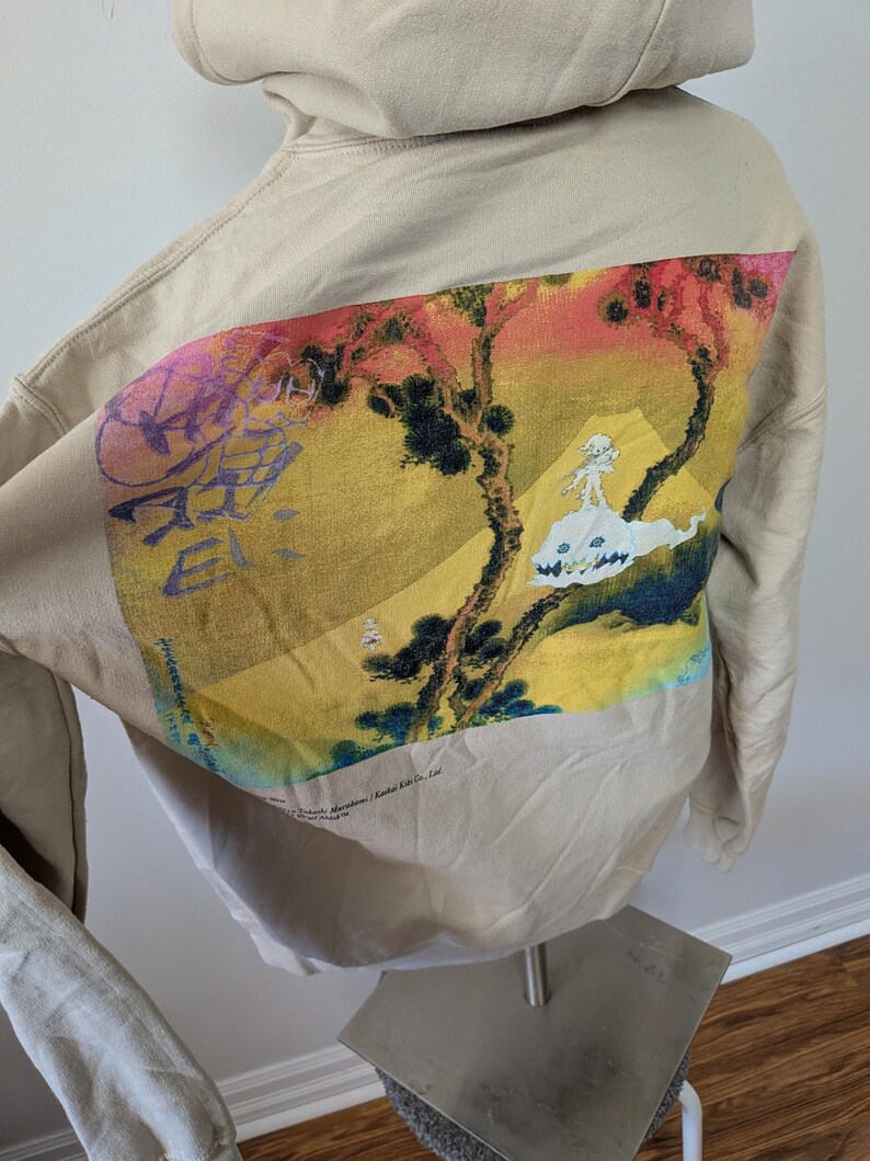 May include: A beige hooded sweatshirt featuring a large rectangular graphic on the back. The print displays a stylized landscape with yellow, red, and green tones, a white creature, and purple text. The text "Takashi Murakami / Kaikai Kiki Co., Ltd." is visible.
