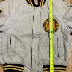 May include: A gray bomber jacket with black and yellow striped ribbing at the collar, cuffs, and waistband. The jacket has a zipper closure and a patch on the left chest that reads "Steelworkers of America United."