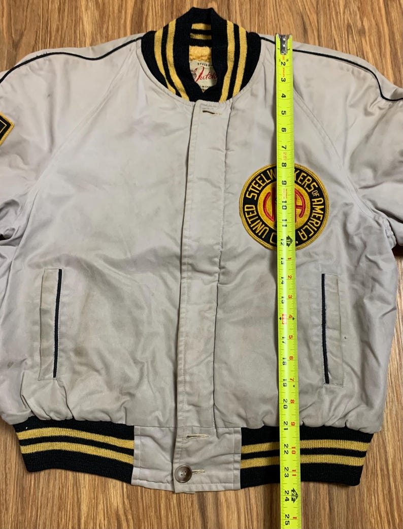 May include: A gray bomber jacket with black and yellow striped ribbing at the collar, cuffs, and waistband. The jacket has a zipper closure and a patch on the left chest that reads "Steelworkers of America United."