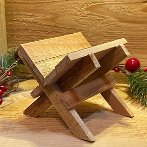 May include: A wooden manger, a traditional nativity scene prop, made of natural wood with a rustic finish. The manger is a simple, rectangular structure with a slightly angled top.