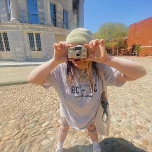 May include: A person wearing a light green beanie and a grey t-shirt with the text "AC/DC Raider Nation" printed on it. They are holding a silver camera in front of their face and standing on a cobblestone street.