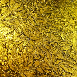May include: Close-up of a textured surface with a repeating floral pattern. The design is embossed and features a metallic gold finish with darker shadows, creating a sense of depth and dimension. The overall effect is opulent and decorative.