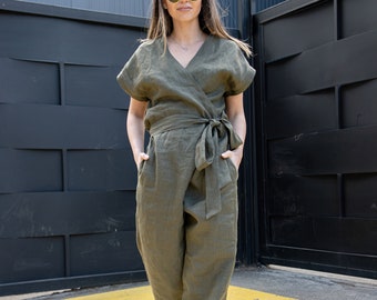 May include: A woman wearing a green linen jumpsuit with a wrap style top and a tie belt. She is wearing black combat boots and round sunglasses.