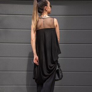 K&ouml;nnte beinhalten: Ein schwarzes &auml;rmelloses Oberteil mit einem durchsichtigen Mesh-Einsatz im R&uuml;cken. Das Oberteil ist drapierend und an der Taille gerafft. Das Model tr&auml;gt eine schwarze Hose und wei&szlig;e Sneaker.