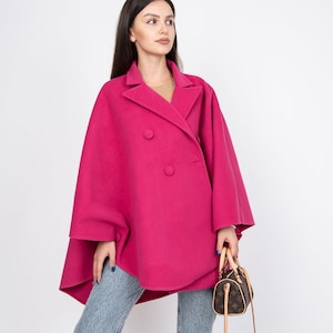 May include: A woman wearing a pink double-breasted coat with a brown handbag. The coat has a wide, flowing silhouette and is made of a soft, textured fabric.