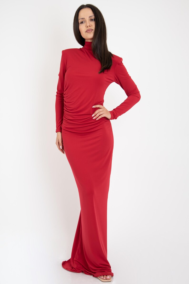 May include: A long-sleeved, red maxi dress with a high neck and a fitted silhouette. The dress has a slight ruffle detail at the shoulders and a gathered skirt.