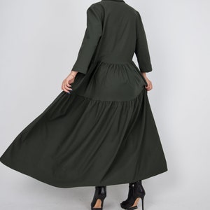 May include: A long, dark green tiered dress with a collared neckline and long sleeves. The dress is flowing and has a relaxed fit. The model is wearing black heeled boots.