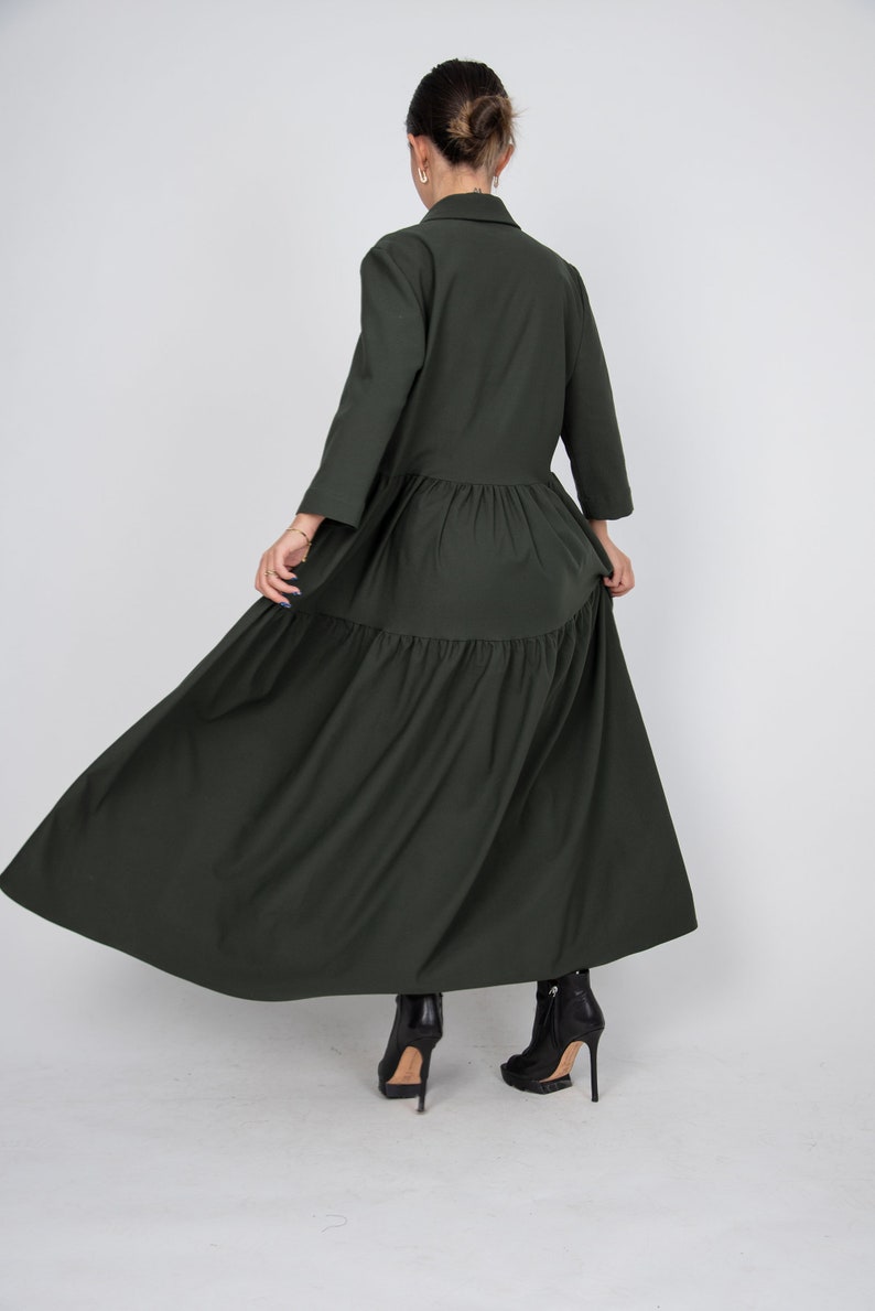 May include: A long, dark green tiered dress with a collared neckline and long sleeves. The dress is flowing and has a relaxed fit. The model is wearing black heeled boots.