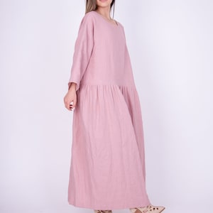 May include: A long, flowing pink linen dress with a loose fit and 3/4 sleeves. The dress has a gathered skirt and is worn by a person with long brown hair.