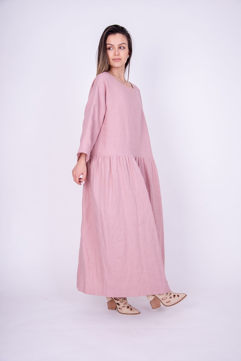 May include: A long, flowing pink linen dress with a loose fit and 3/4 sleeves. The dress has a gathered skirt and is worn by a person with long brown hair.