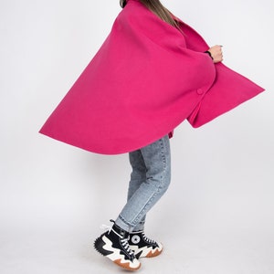 May include: A woman wearing a bright pink cape with a single button closure. She is wearing blue jeans and black and white high-top sneakers.