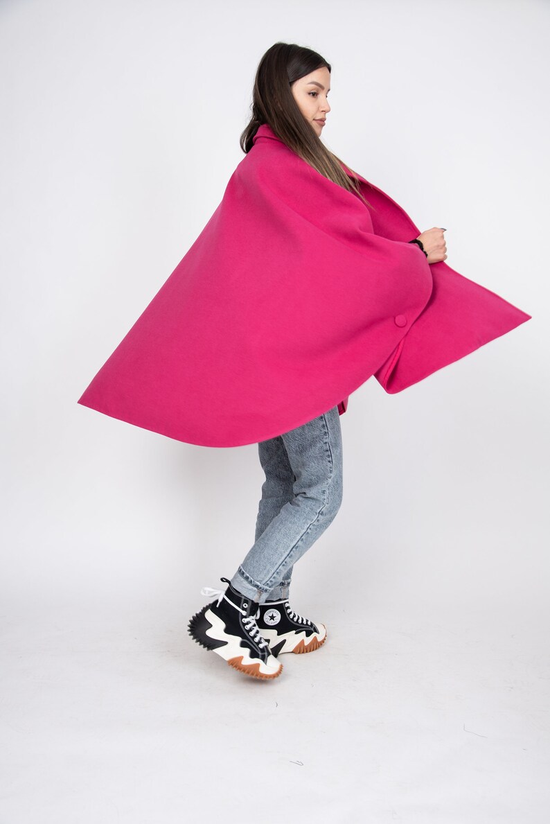 May include: A woman wearing a bright pink cape with a single button closure. She is wearing blue jeans and black and white high-top sneakers.