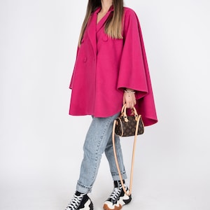 May include: A woman wearing a pink double-breasted cape coat, blue jeans, and black and white high-top sneakers. She is carrying a brown leather handbag with a long strap.