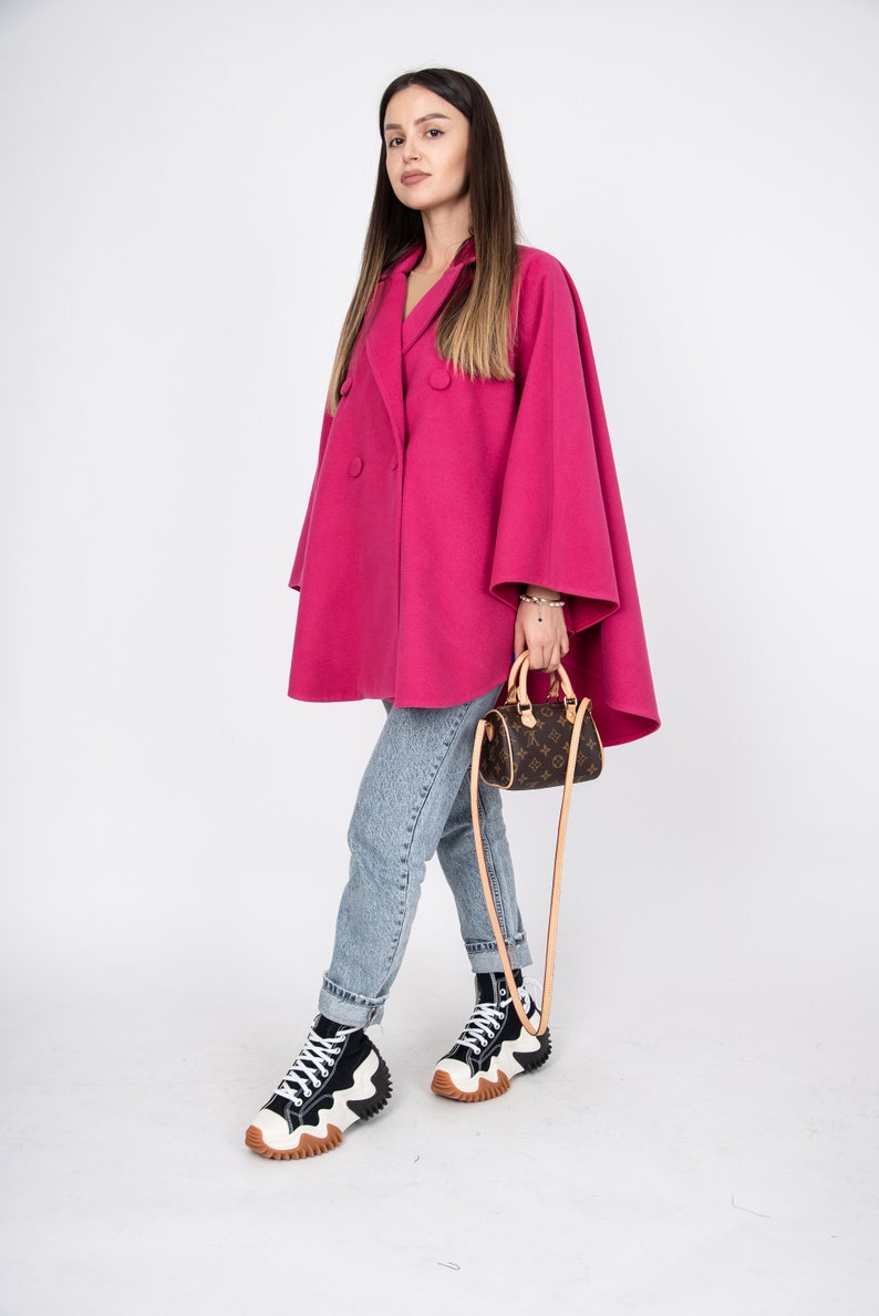 May include: A woman wearing a pink double-breasted cape coat, blue jeans, and black and white high-top sneakers. She is carrying a brown leather handbag with a long strap.