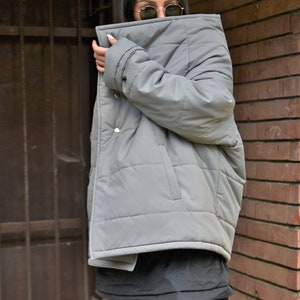 Peut inclure: Une veste matelass&eacute;e grise avec un col montant et une fermeture &eacute;clair. La veste est port&eacute;e par une personne aux cheveux fonc&eacute;s et aux lunettes de soleil.