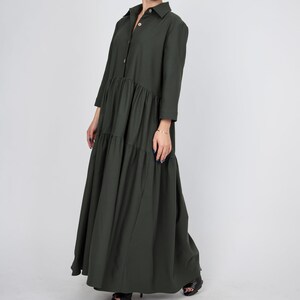 May include: A long, dark green, tiered dress with a button-up collar and long sleeves. The dress has a relaxed fit and falls to the ankles.