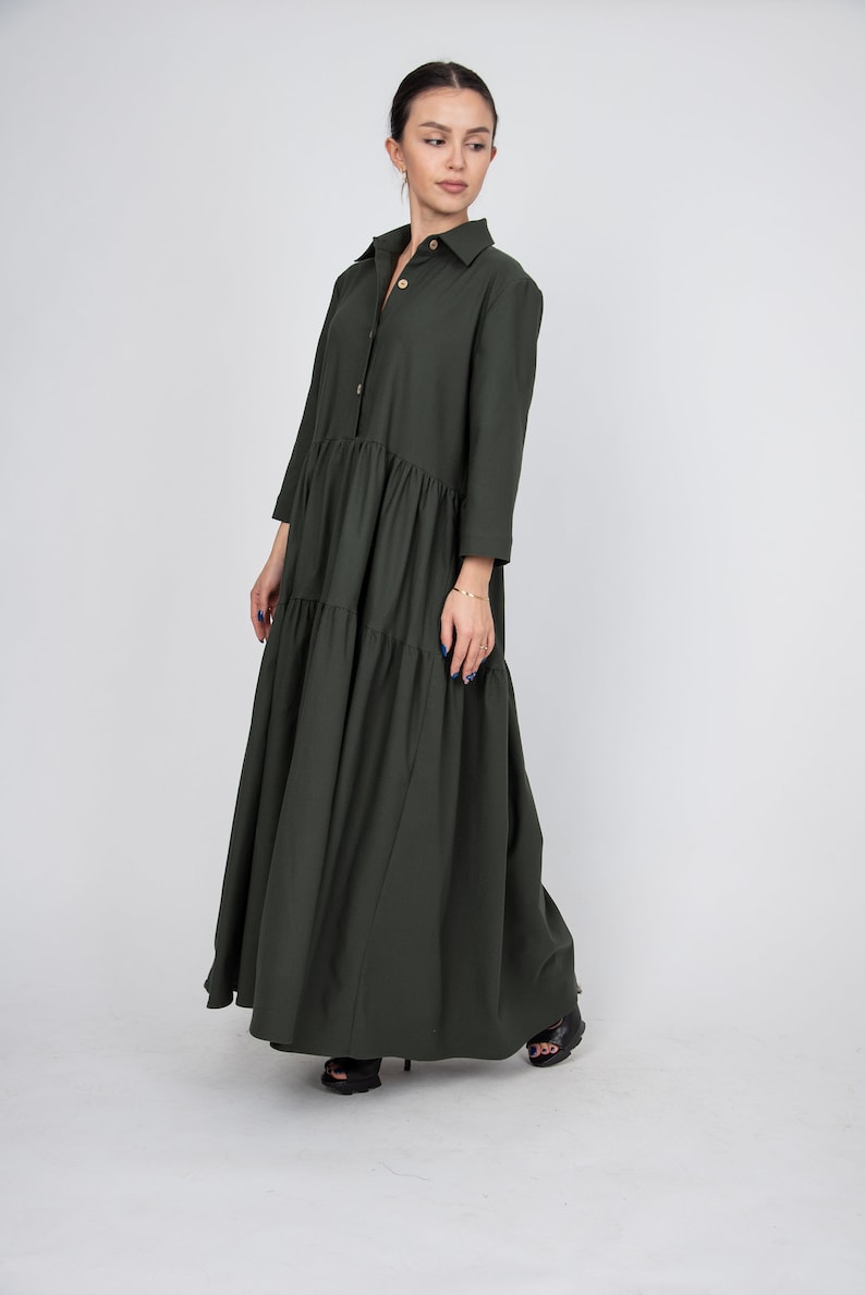 May include: A long, dark green, tiered dress with a button-up collar and long sleeves. The dress has a relaxed fit and falls to the ankles.