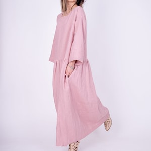 May include: A long, flowing pink linen dress with a relaxed fit and three-quarter sleeves. The dress has a gathered waist and a single pocket on the left side.