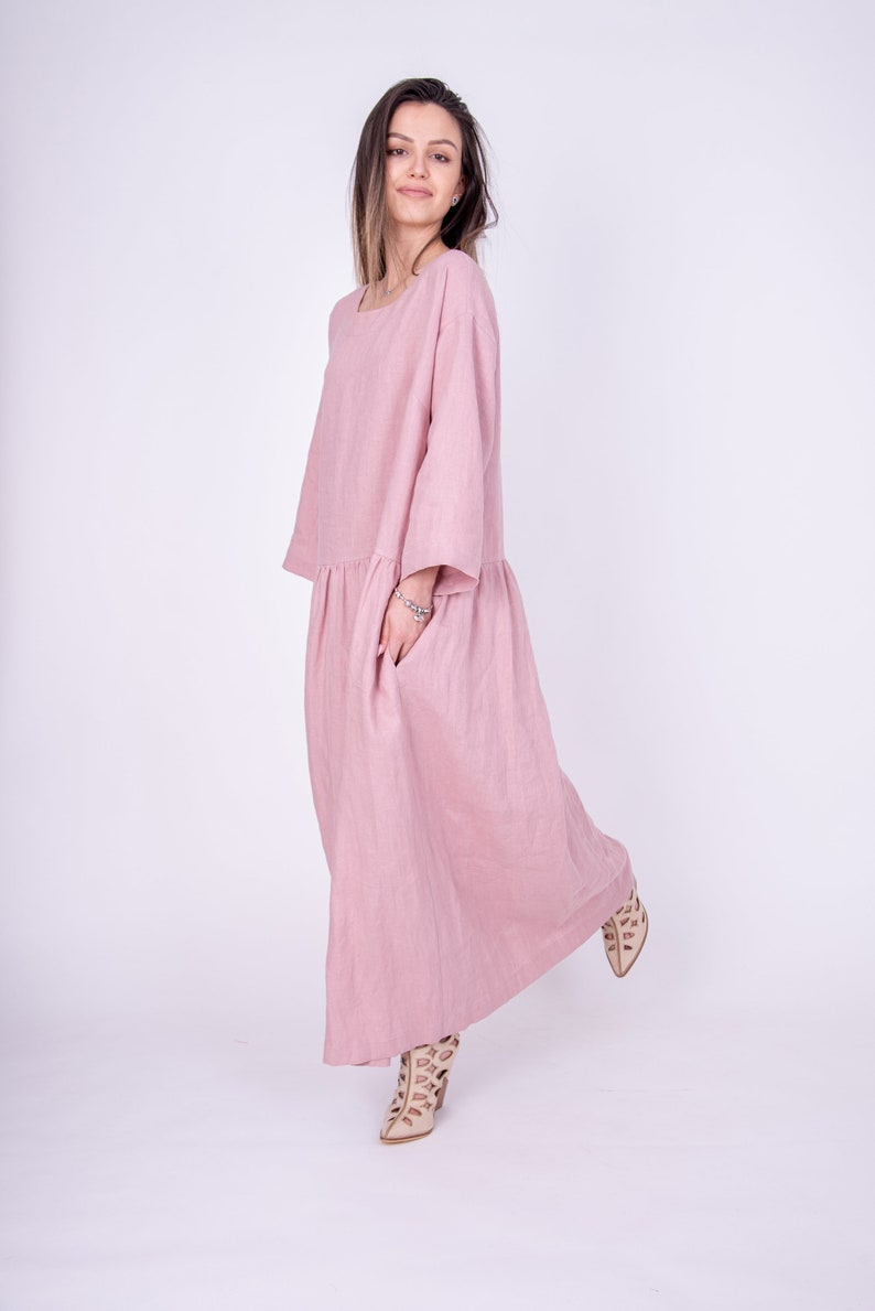 May include: A long, flowing pink linen dress with a relaxed fit and three-quarter sleeves. The dress has a gathered waist and a single pocket on the left side.