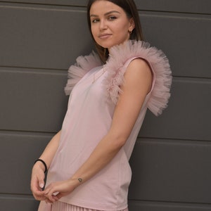 May include: A pink sleeveless top with a ruffled tulle overlay on the shoulders. The top is paired with a pleated skirt in the same color.