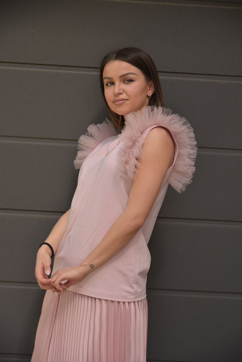 May include: A pink sleeveless top with a ruffled tulle overlay on the shoulders. The top is paired with a pleated skirt in the same color.