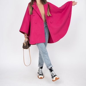 May include: A woman wearing a pink double-breasted coat with large sleeves, blue jeans, and white sneakers. She is carrying a small brown leather handbag.