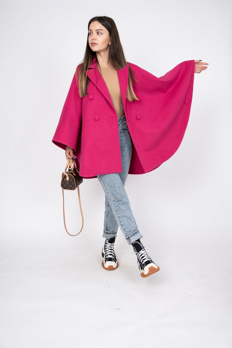 May include: A woman wearing a pink double-breasted coat with large sleeves, blue jeans, and white sneakers. She is carrying a small brown leather handbag.