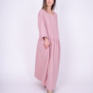 May include: A woman wearing a long, pink linen dress with a gathered skirt. She is standing on a white background.