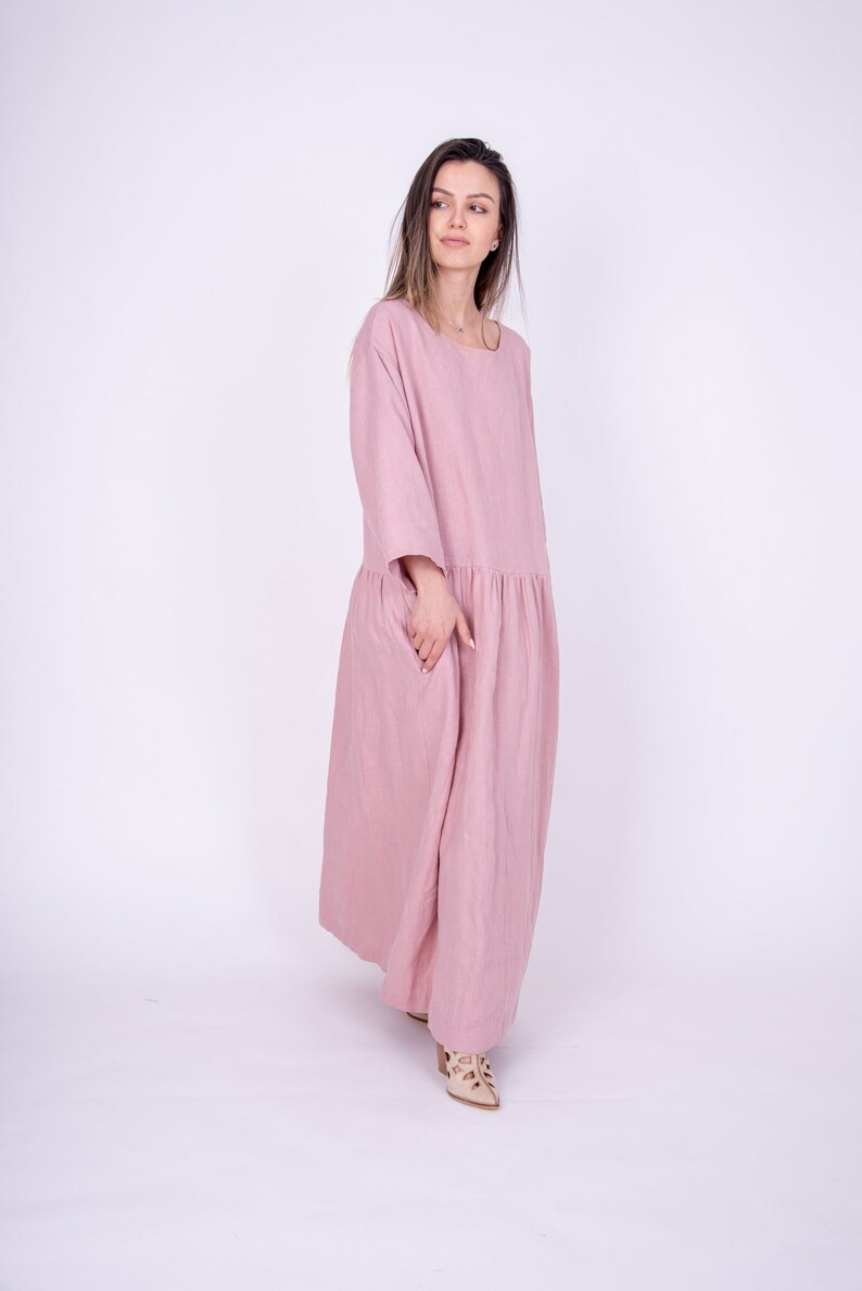 May include: A woman wearing a long, pink linen dress with a gathered skirt. She is standing on a white background.