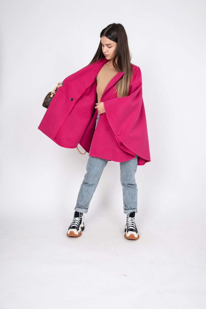 May include: A woman wearing a pink cape with a single button closure, blue jeans, and black and white sneakers. She is holding a brown handbag.