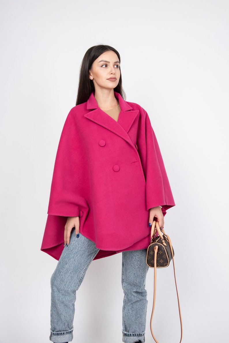May include: A woman wearing a pink double-breasted coat with a brown handbag. The coat has a wide, flowing silhouette and is made of a soft, textured fabric.