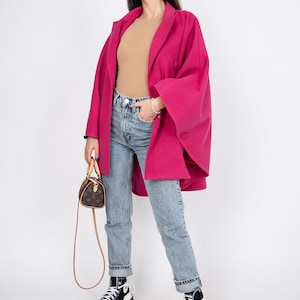 May include: A woman wearing a pink coat, blue jeans, and black and white high-top sneakers. She is carrying a small brown handbag with a strap.