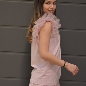 May include: A pink sleeveless top with a ruffled tulle overlay on the shoulders. The top is paired with a pleated pink skirt.
