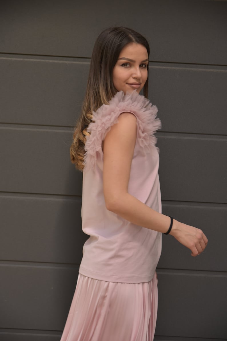 May include: A pink sleeveless top with a ruffled tulle overlay on the shoulders. The top is paired with a pleated pink skirt.