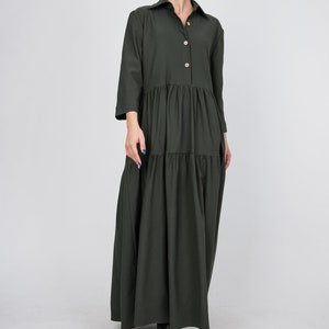 May include: A long, dark green, tiered dress with a button-up collar and long sleeves. The dress has a relaxed fit and falls to the ankles.